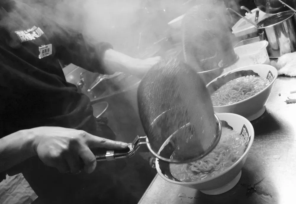 湯気の立つ厨房で、湯切りした麺をらーめんの丼に入れる調理風景